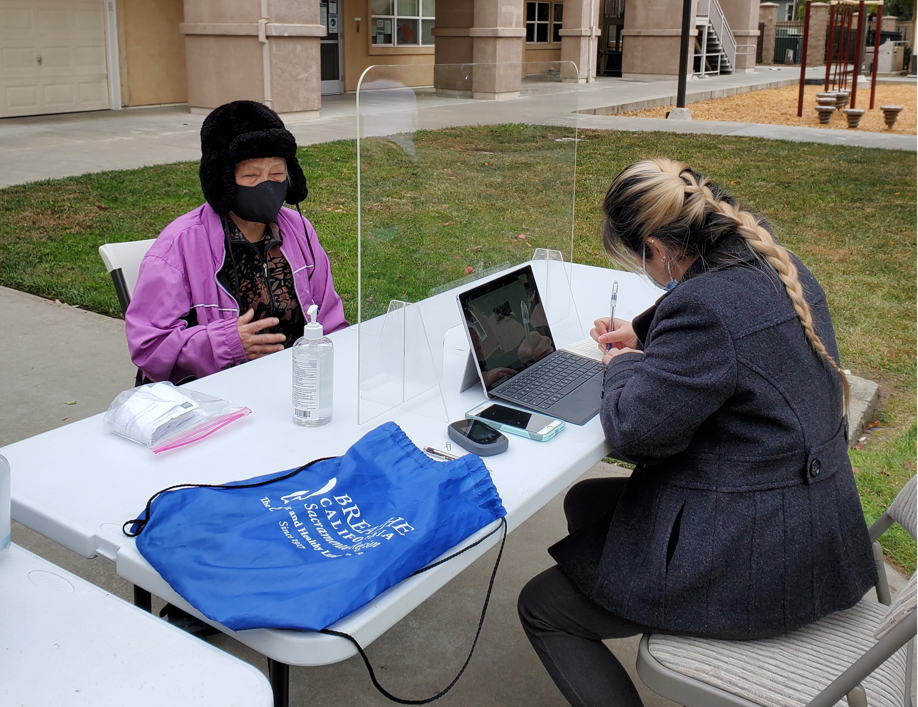 Carshare site outdoor outreach events, two women talking at table with plexi glass barrier. Women are wearing face coverings as a COVID precaution. 