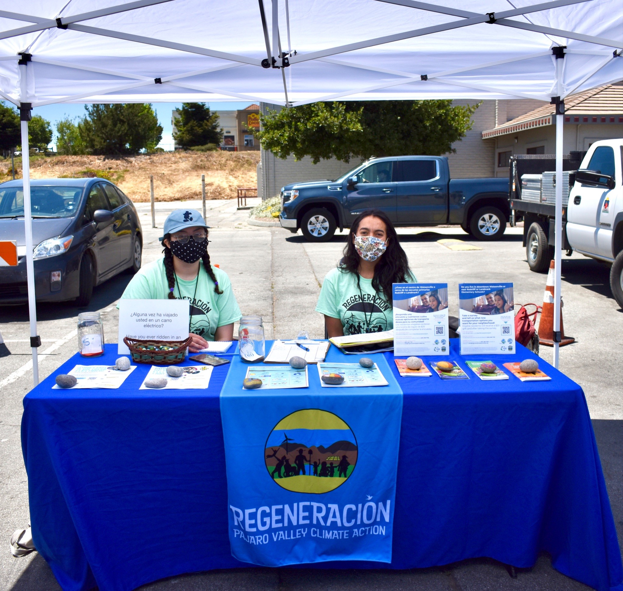 Social Good Fund Project Regeneration survey team members tabling at Farmers’ Market in Watsonville. The two pictured are sitting at an outdoor booth with materials displayed on the table.