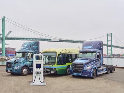 Two zero-emission trucks and a zero emission bus next to charger and with port backdrop