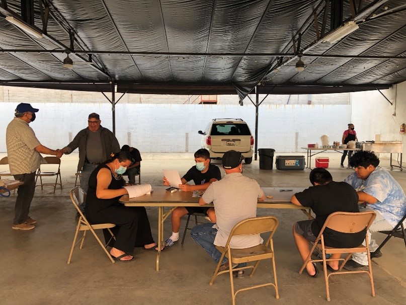 Several masked residents gather around a table at an indoor outreach event wearing masks to protect against COVID-19