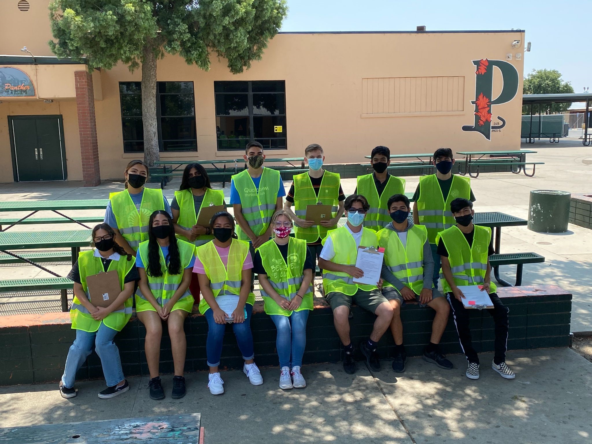 Student interns completing an infrastructure walk audit of pedestrian safety measures at Olive Elementary. 13 students wearing safety vests and COVID masks pose in front of school building.