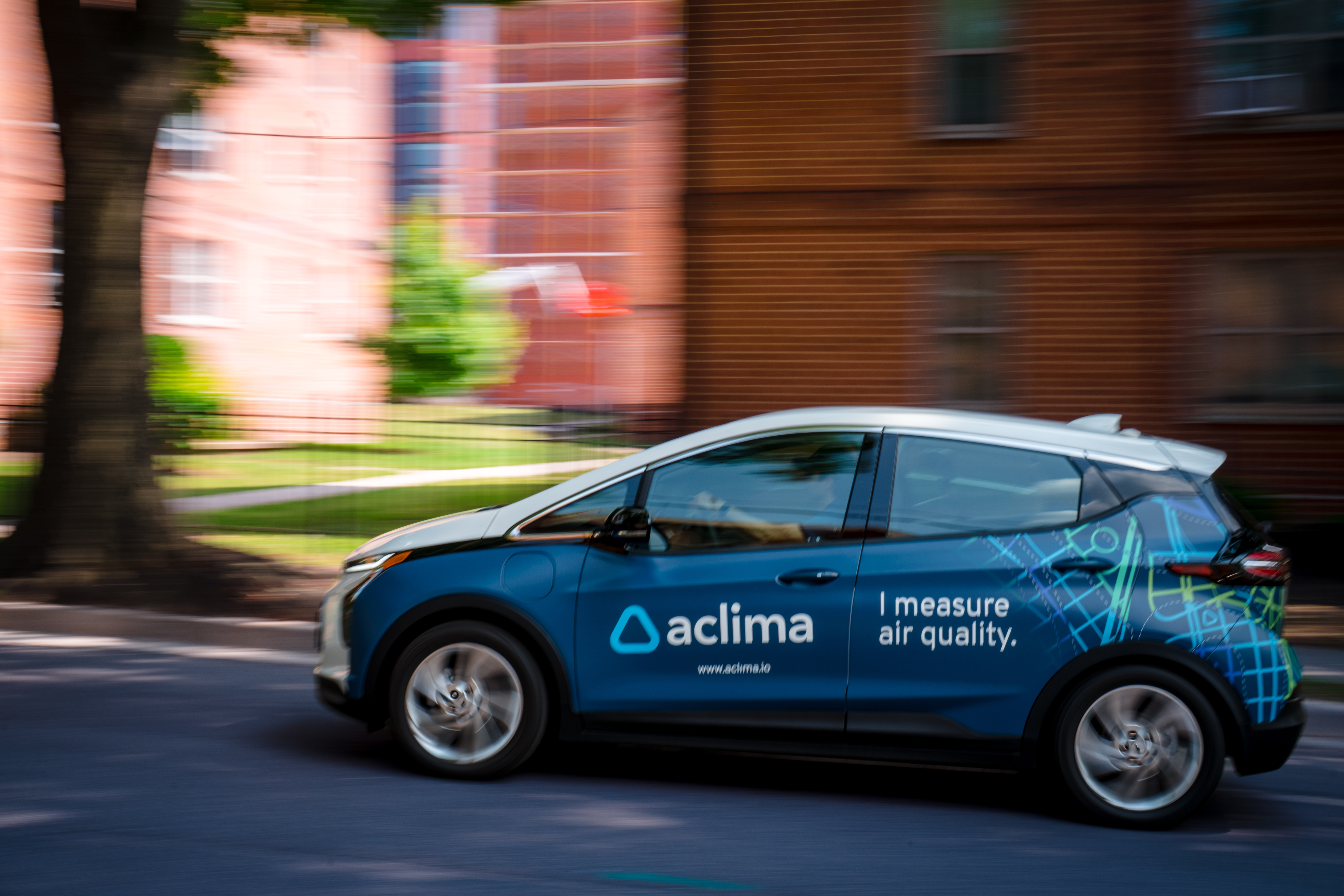 Sedan driving on street with blurred buildings in background. Vehicle has a blue wrap with the Aclima, Inc. logo and text that reads, "I measure air quality."