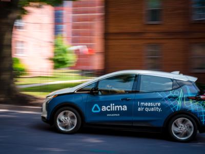 Small sedan driving on street with blurred buildings in the background. Vehicle has a blue wrap with the Aclima, Inc. logo, and text that reads "I measure air quality."